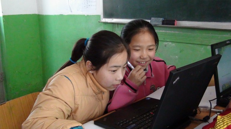 Two female Chinese students looking at a laptop in a classroom with chipped paint on the walls.