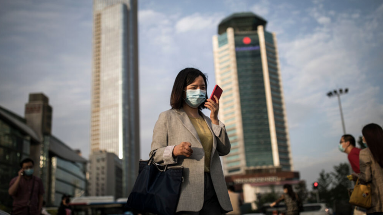 A professional looks at their phone while wearing a mask in a busy downtown area.