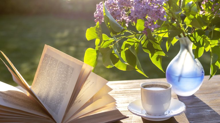 Little white cup of espresso coffee, opened book, blue semi-transparent vase with purple lilac flowers on rustic wooden table in the garden at spring morning after sunrise or at evening before sunset