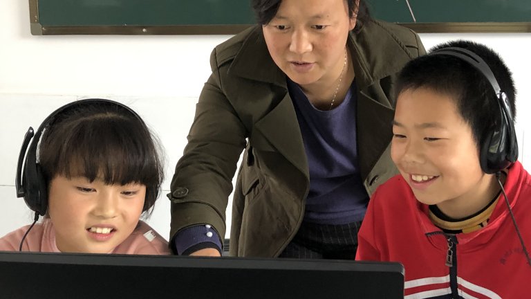 Two smiling Chinese students wearing headphones and looking at a computer screen with a teacher behind them gesturing to the screen.