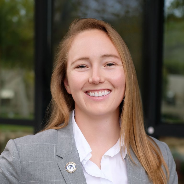 A headshot of Lyndsea Warkenthien. Lyndsea is smiling and wearing a grey blazer and white shirt.