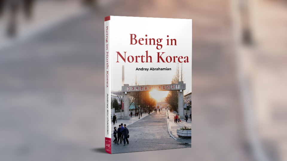 Cover of the book 'Being in North Korea' by Andray Abrahamian on a blurred background of a road, a cropped scene from the cover image