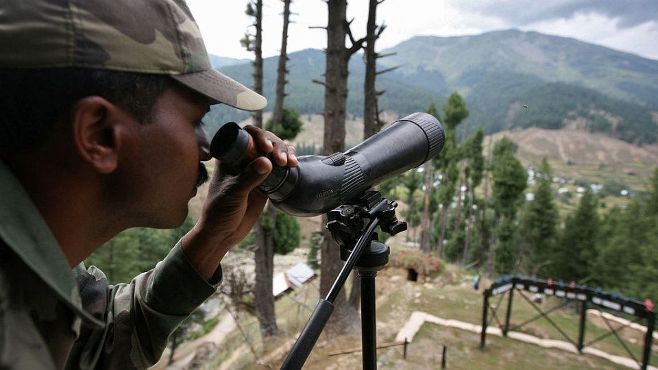 An Indian Army soldier looking through a military monocular over hills in the background