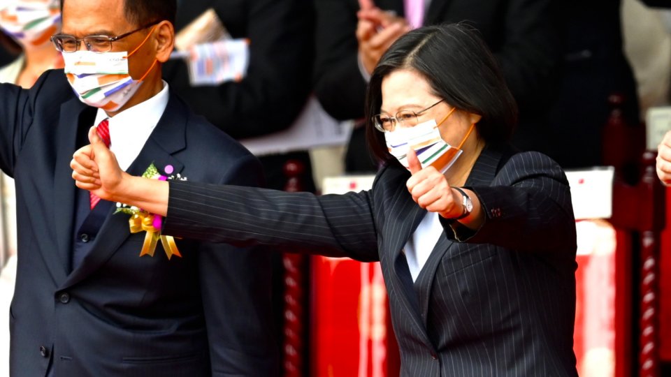 An Asian woman waring a face mask standing among a crowd gesturing with her hands