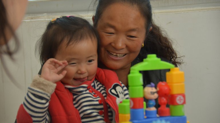 Toddler sitting on mom's lap looking at a colorful plastic building block structure.