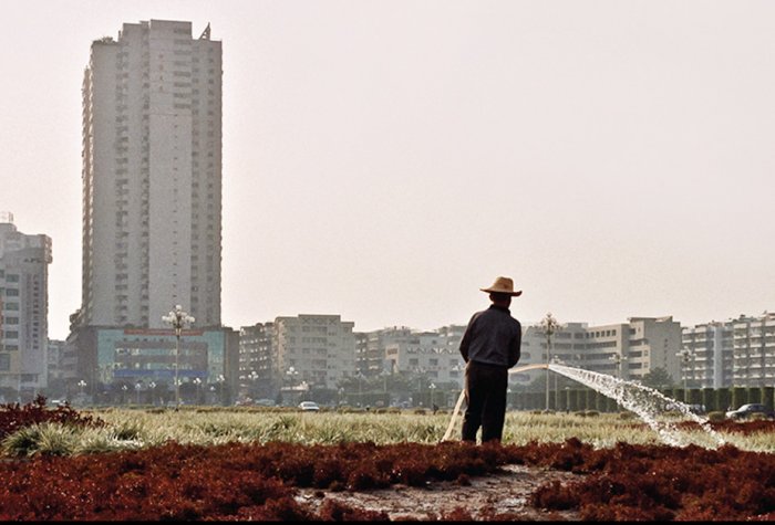 Farmer watering fields in front of a cityscape.