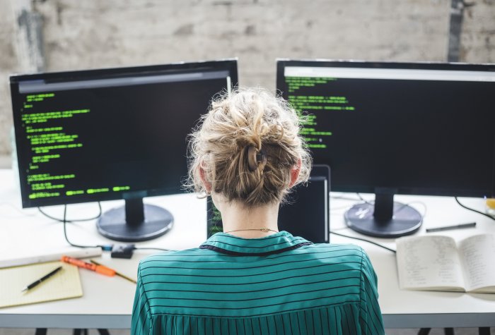 Female computer coder looking at a dual monitor with code on it.
