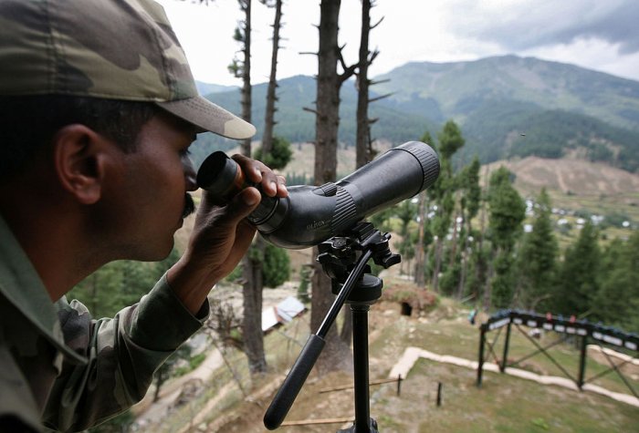 An Indian Army soldier looking through a military monocular over hills in the background
