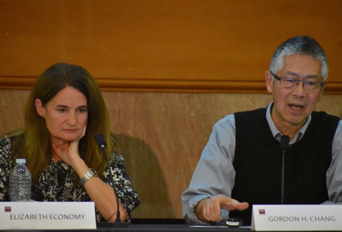 A man and a woman seated at a panel table, the man speaking