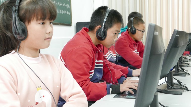 Three young students wearing headphones and working on desktop computers in a classroom.