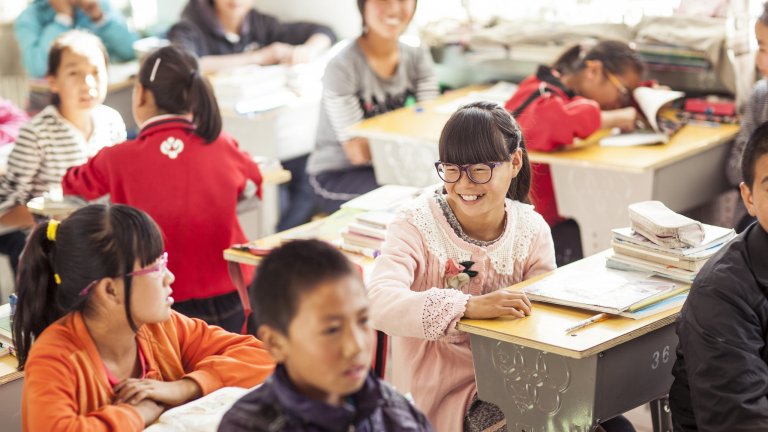 Middle school kids at school desks smiling at each other while wearing glasses.
