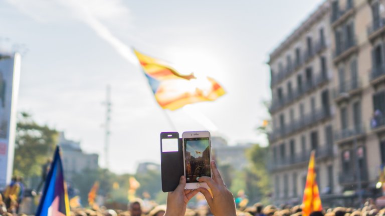 Mobile phone, held in hands, taking a photo of a street with crowds carrying flags. Mobile phone, held in hands, taking a photo of a street with crowds carrying flags.